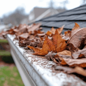 Close-up view of a home’s gutter clogged with brown and red maple leaves during fall, showing how seasonal debris can block water flow along the roof edge.